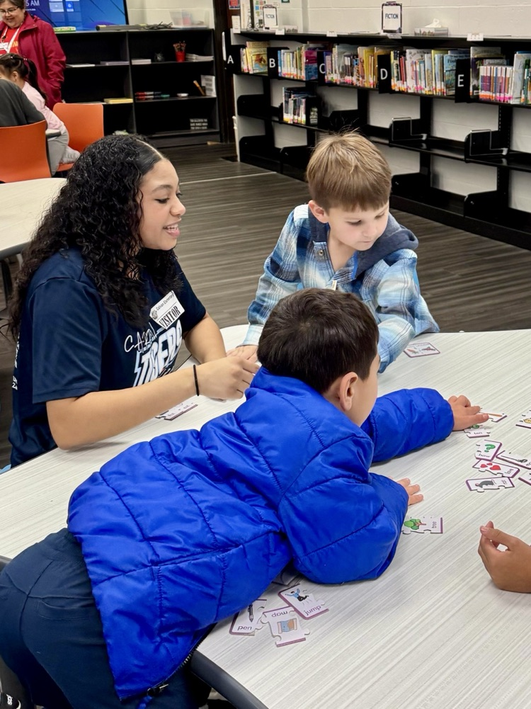 Elementary students sit at tables in a school library working on reading activities.