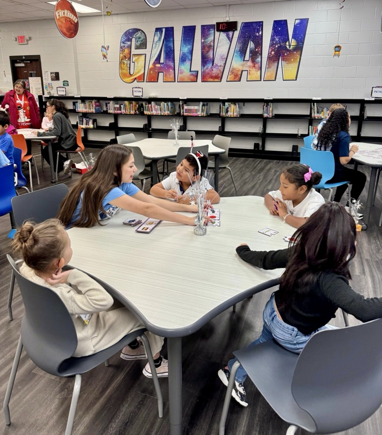 Elementary students sit at tables in a school library working on reading activities.