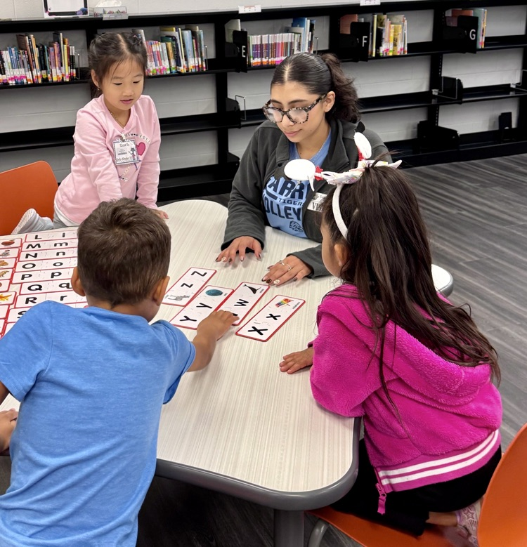 Elementary students sit at tables in a school library working on reading activities.