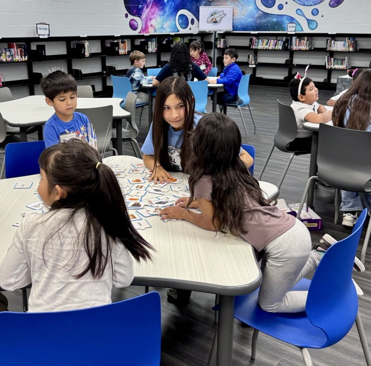 Elementary students sit at tables in a school library working on reading activities.