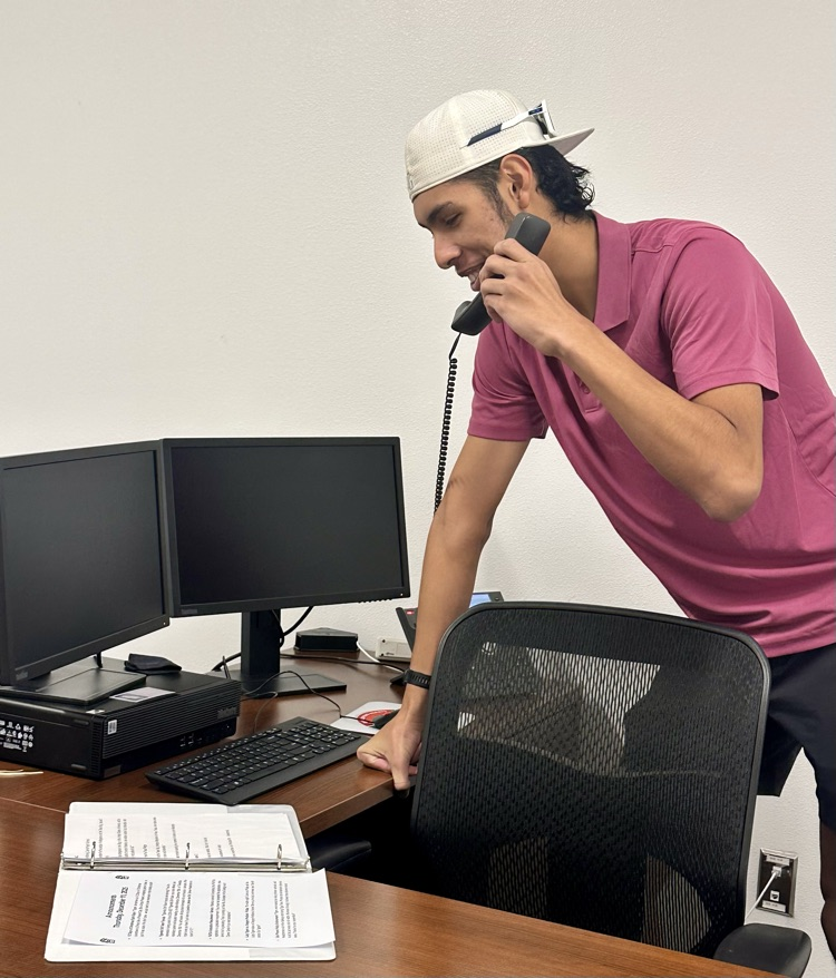 Alt Text: Two students read morning announcements into the intercom phone while standing at a desk with computers and papers.