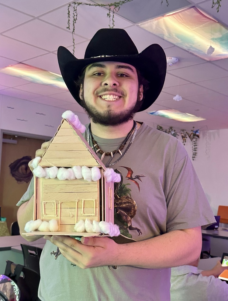 Alt Text: A student smiles while holding a cabin decorated with cotton snow and close-up of small Christmas cabin model on table,