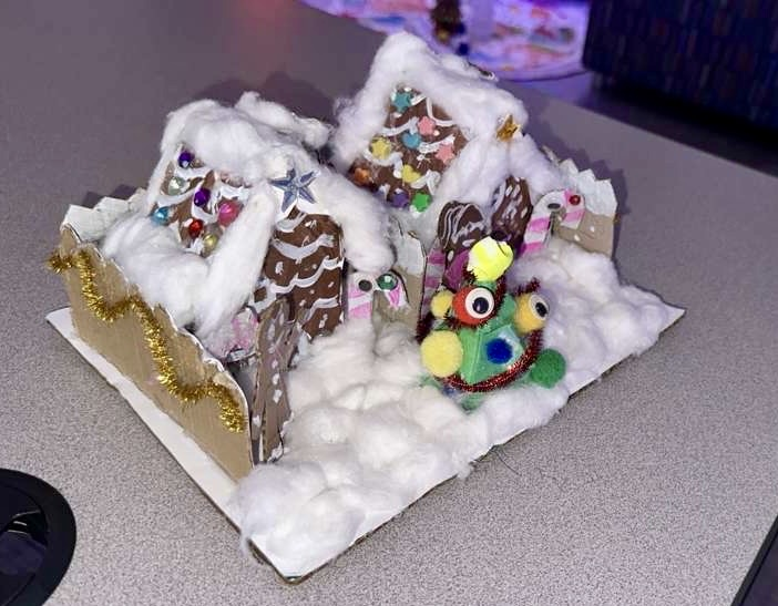 Alt Text: A student smiles while holding a cabin decorated with cotton snow and close-up of small Christmas cabin model on table,