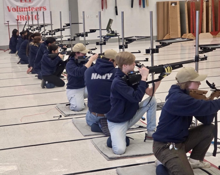 Alt Text: Carroll High School NJROTC cadets kneel in a straight line on mats inside a gym, aiming rifles at targets during a marksmanship competition