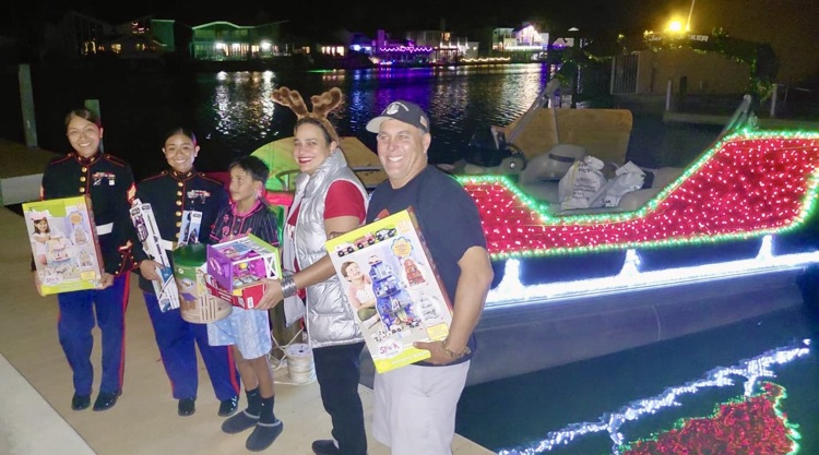 Alt Text: Students and adults stand together on a dock at night holding donated toys during a holiday toy drive,