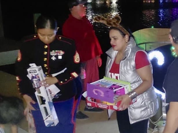 Alt Text: Students and adults stand together on a dock at night holding donated toys during a holiday toy drive,