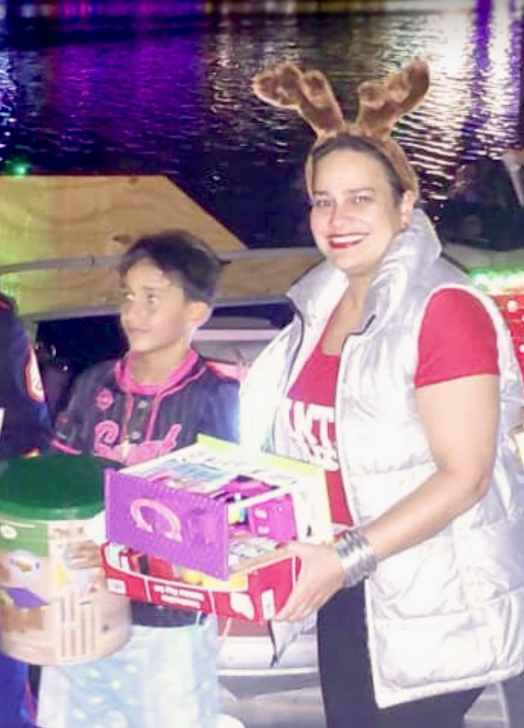 Alt Text: Students and adults stand together on a dock at night holding donated toys during a holiday toy drive,