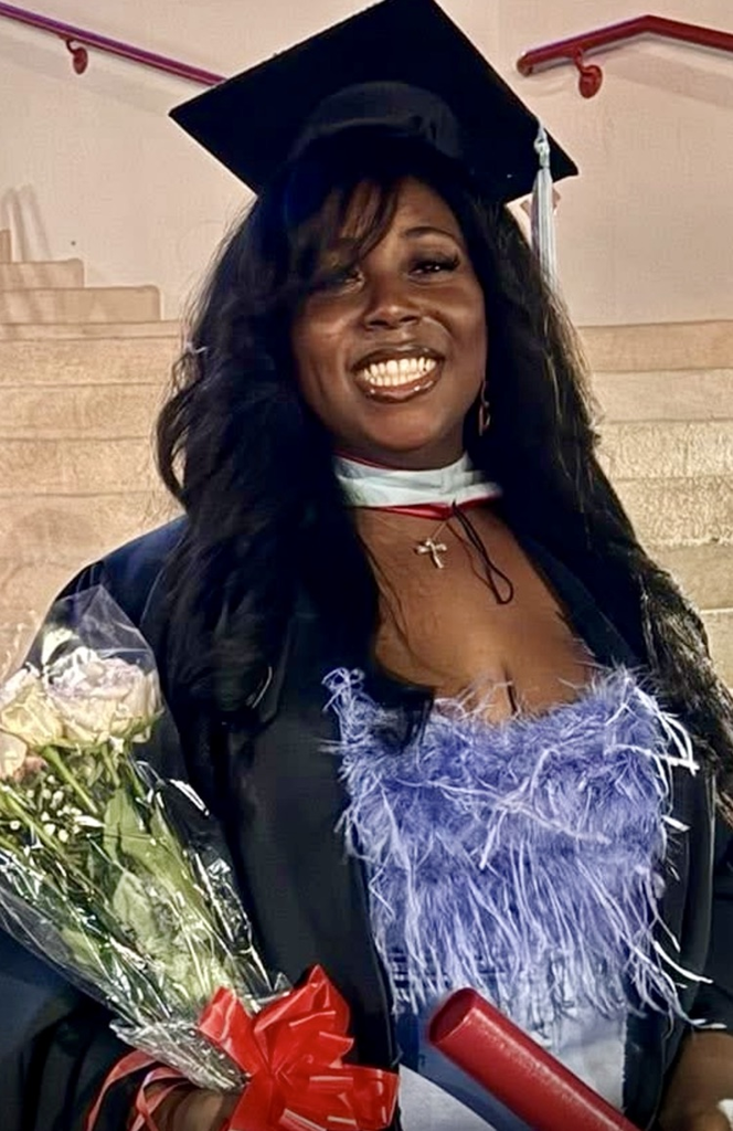 Alt Text: A smiling teacher wearing a graduation cap and gown holds a bouquet of flowers