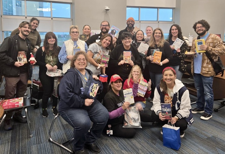 ⸻  Alt Text – Image 1: Large group of smiling staff members in a school library holding books and holiday gift items during a festive Hug in a Mug and book exchange event.