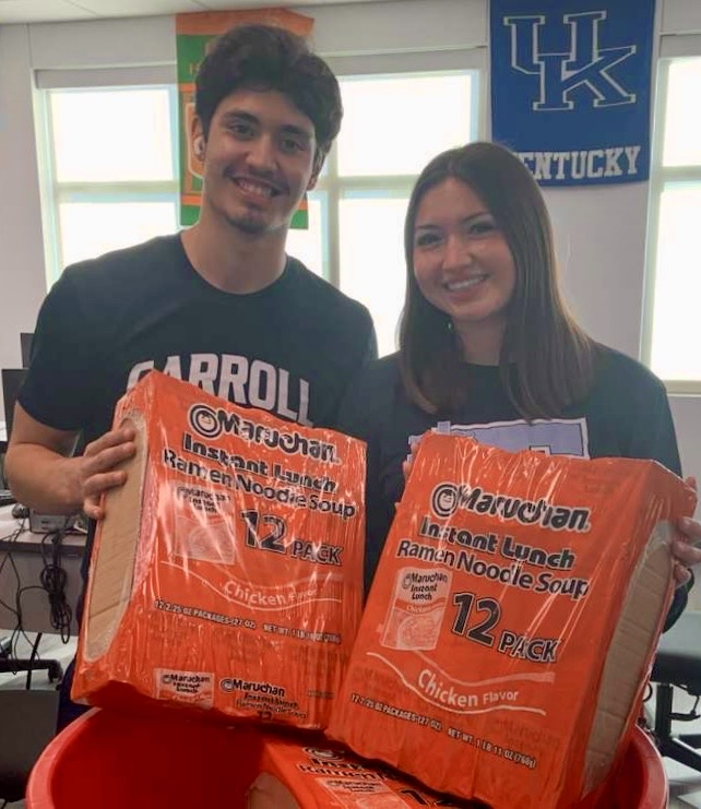 Group of NHS students standing behind red food donation bins labeled for a food bank, holding donated food items and smiling at the camera. 
