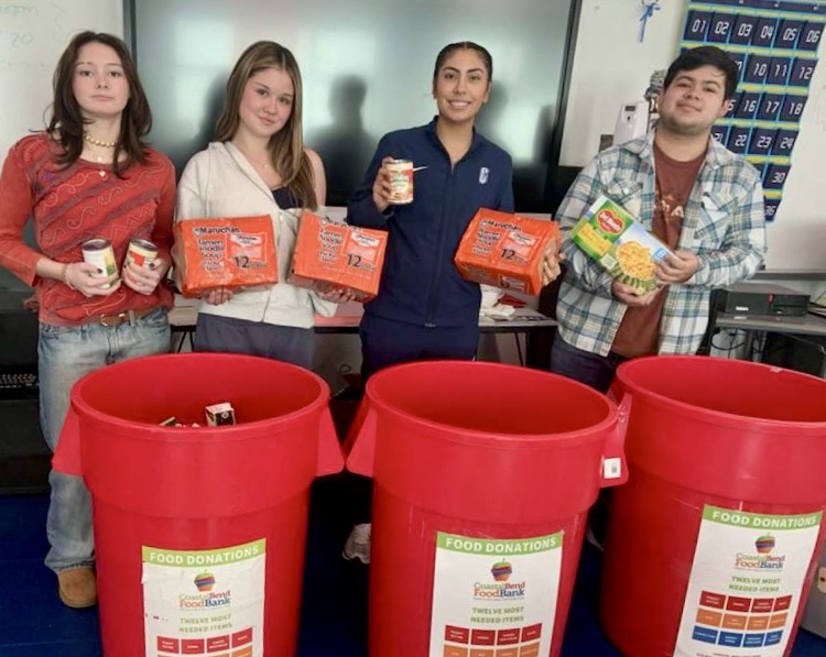 Group of NHS students standing behind red food donation bins labeled for a food bank, holding donated food items and smiling at the camera. 