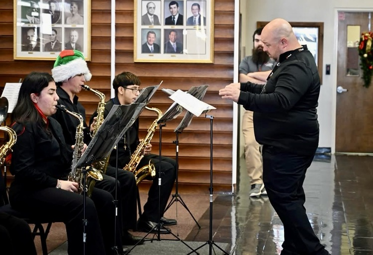 Carroll High School band students perform holiday music inside the district Administration Building, dressed in black concert attire and led by conductor Mr. Sorrows, as staff watch and listen.