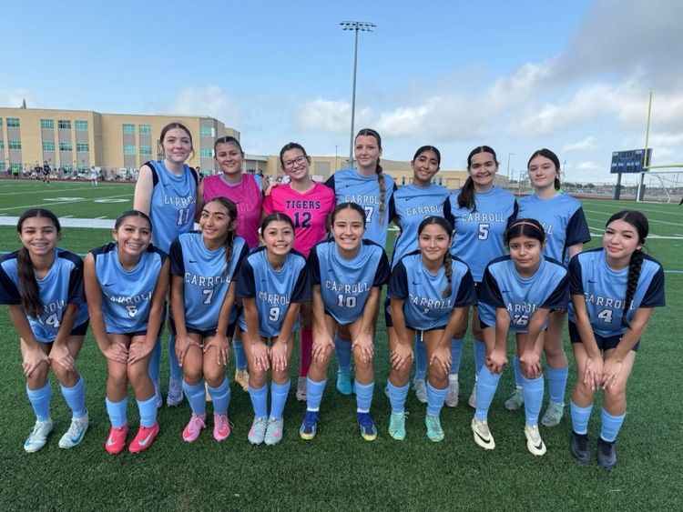 Carroll High School girls soccer team poses together on the field in blue uniforms, smiling and ready for the season, with coaches standing behind them.