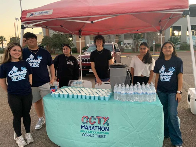 Students volunteering at a hydration station with water cups and bottles under a tent at the CCTX Marathon in Corpus Christi.
