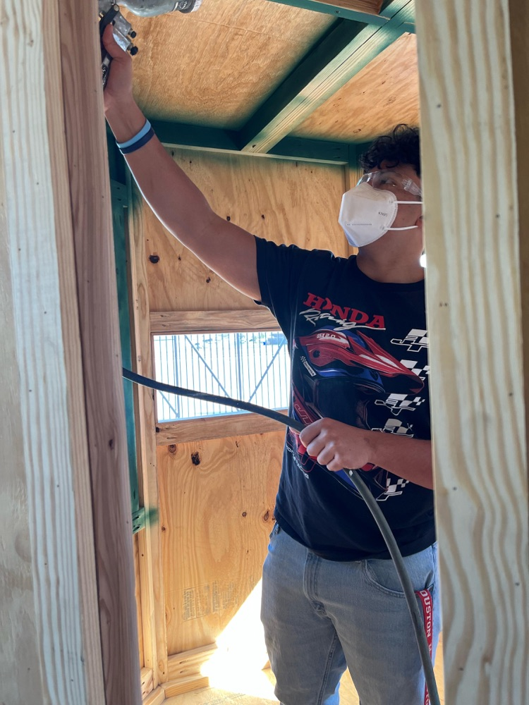 Short Alt Text: Student wearing safety mask spray painting inside wooden structure.