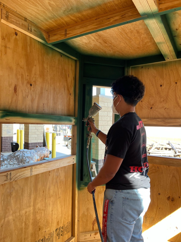 Short Alt Text: Student wearing safety mask spray painting inside wooden structure.