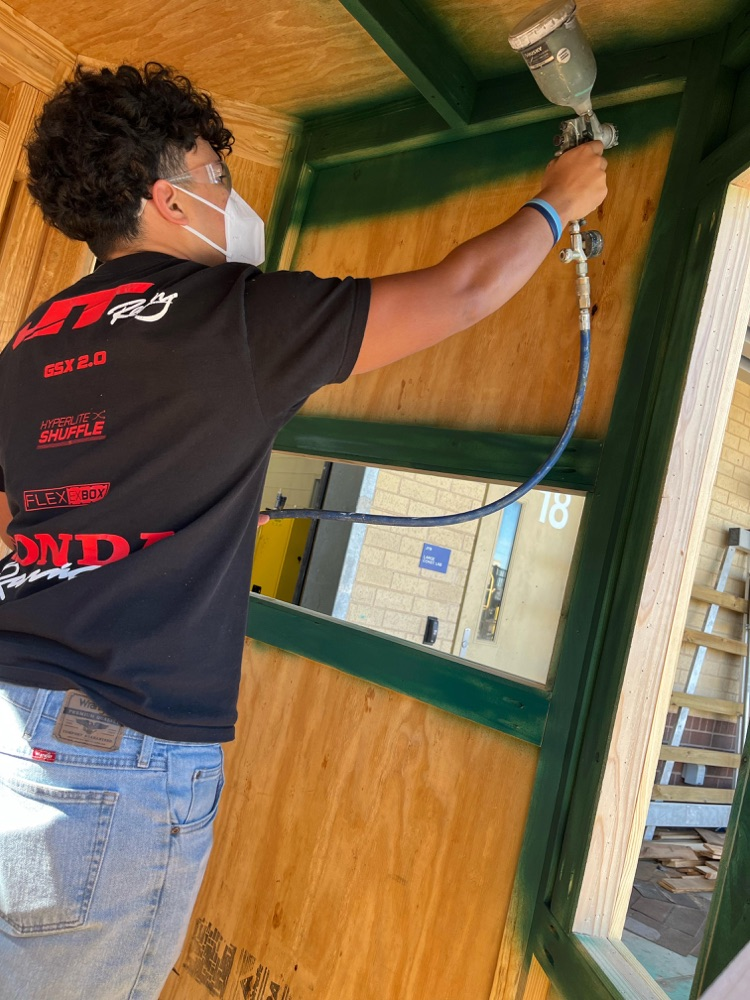 Short Alt Text: Student wearing safety mask spray painting inside wooden structure.