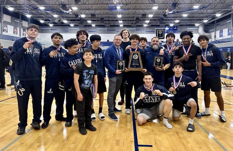 : Carroll High School boys wrestling team poses with medals and district championship plaques on a gym floor after winning the 2025–26 District 15-5A title.