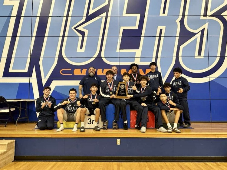: Carroll High School boys wrestling team poses with medals and district championship plaques on a gym floor after winning the 2025–26 District 15-5A title.