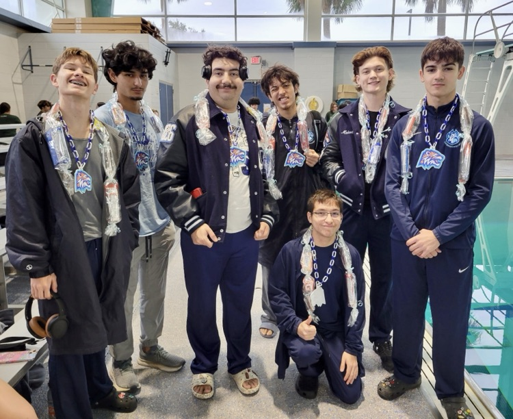 Group of Carroll High School swim seniors standing together on the pool deck wearing team apparel and senior candy leis, smiling and posing for a photo at a swim meet.