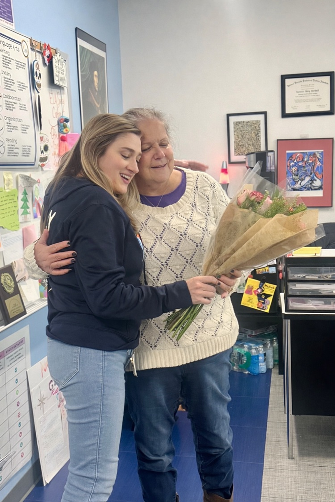 Alt Text: Ms. Sue Marshall stands in her classroom holding a bouquet of flowers and smiling. Another image shows her hugging a colleague while holding the bouquet, with student work and posters visible around the room.