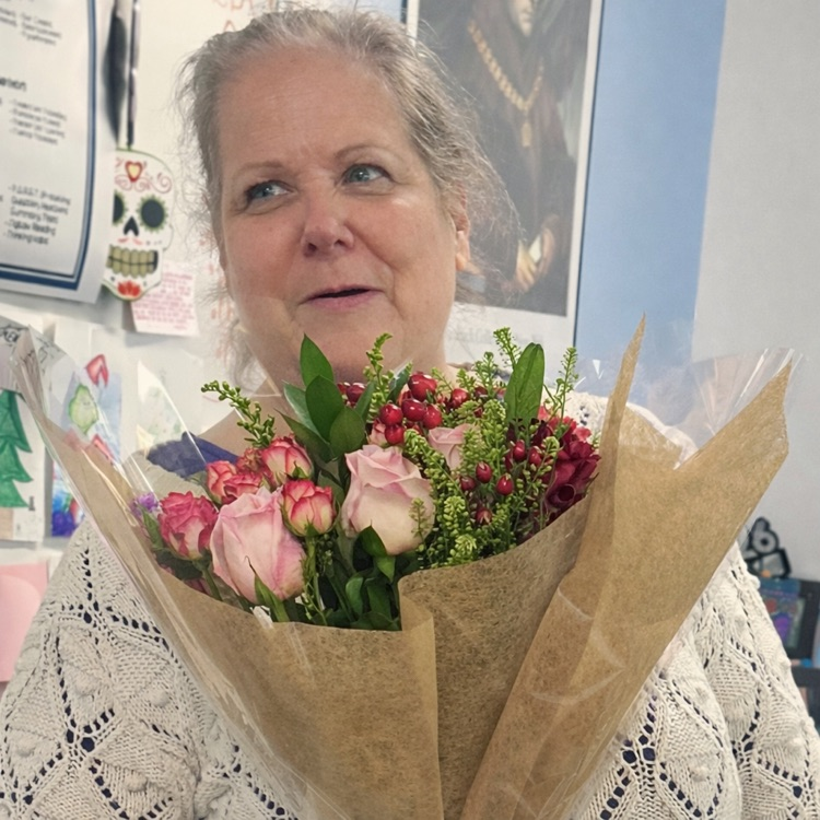 Alt Text: Ms. Sue Marshall stands in her classroom holding a bouquet of flowers and smiling. Another image shows her hugging a colleague while holding the bouquet, with student work and posters visible around the room.