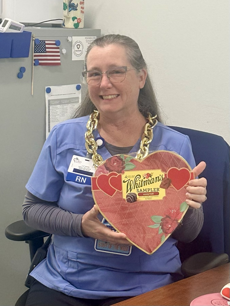 nurse in blue scrubs is smiling and holding a heart-shaped Whitman’s Sampler chocolate box, while another staff member stands beside her 