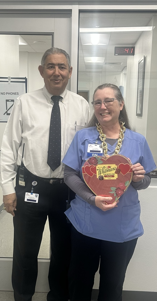 nurse in blue scrubs is smiling and holding a heart-shaped Whitman’s Sampler chocolate box, while another staff member stands beside her 