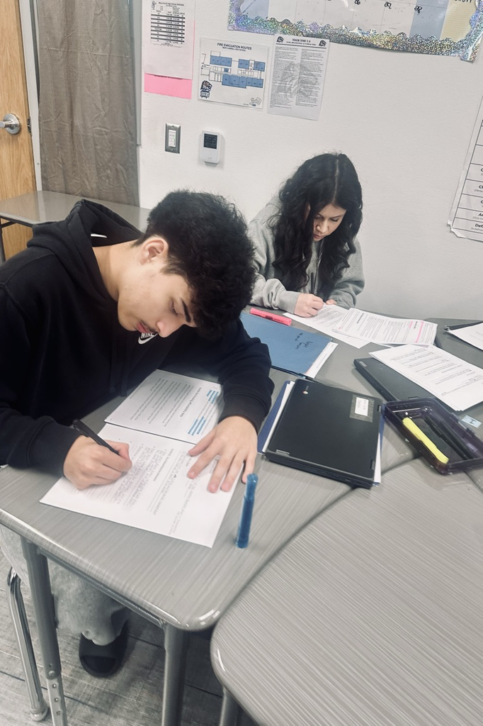 Alt Text: Students seated at desks in an English classroom, focused on reading passages, highlighting text, and writing responses on printed assignments while laptops and notebooks are open on the tables.