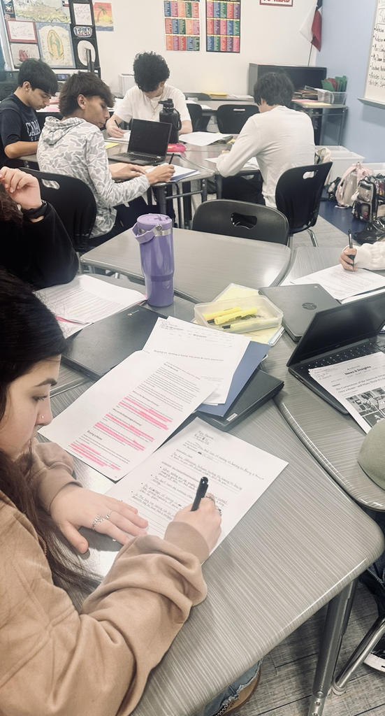 Alt Text: Students seated at desks in an English classroom, focused on reading passages, highlighting text, and writing responses on printed assignments while laptops and notebooks are open on the tables.
