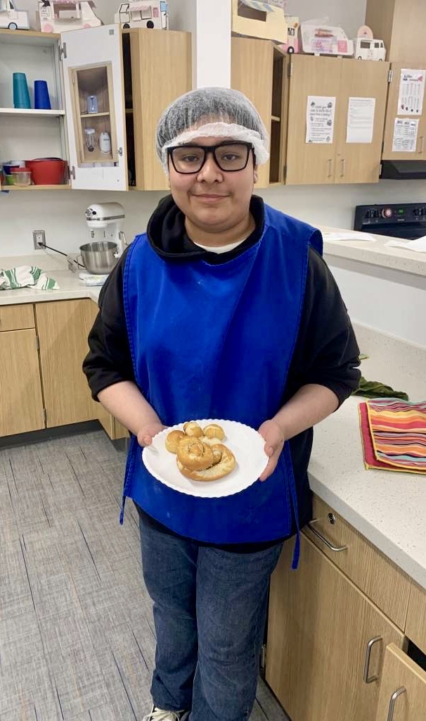 Alt text: Culinary students wearing aprons and hairnets proudly holding plates of freshly made pancakes and soft pretzels in a school kitchen.