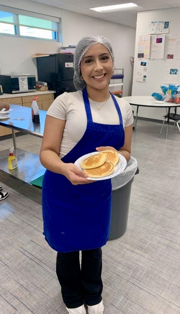 Alt text: Culinary students wearing aprons and hairnets proudly holding plates of freshly made pancakes and soft pretzels in a school kitchen.
