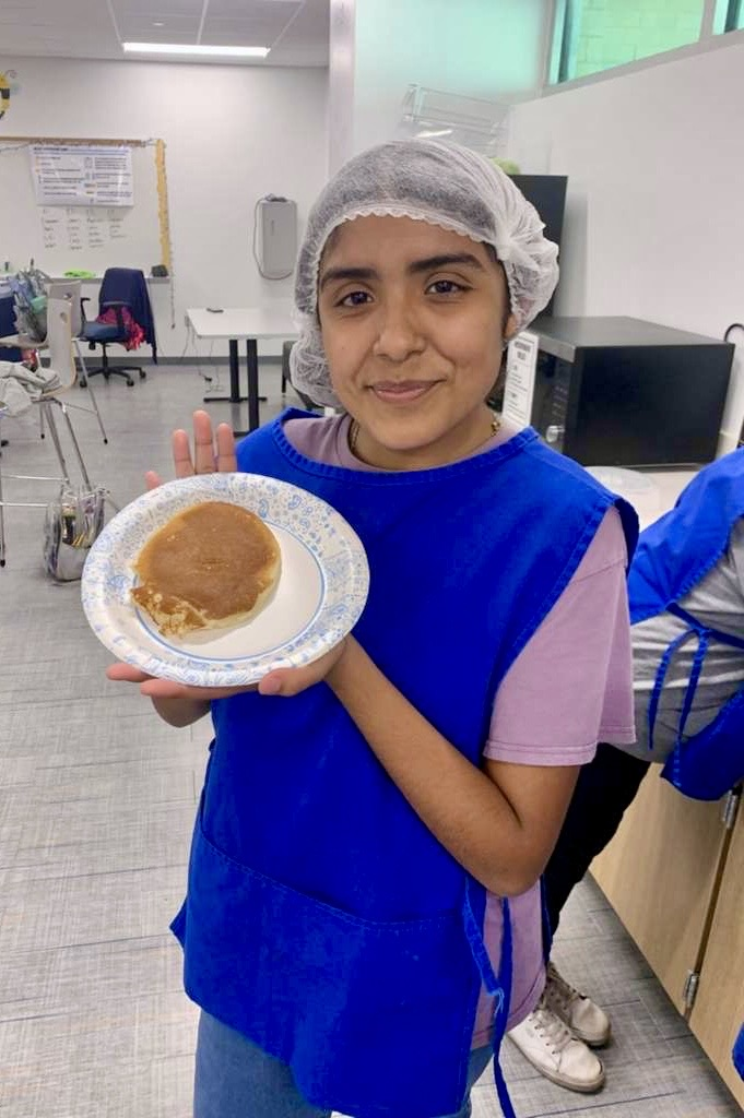 Alt text: Culinary students wearing aprons and hairnets proudly holding plates of freshly made pancakes and soft pretzels in a school kitchen.