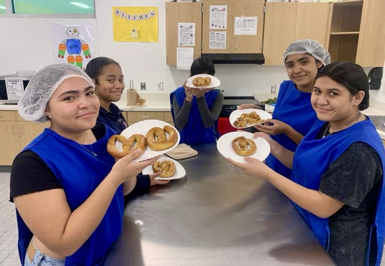Alt text: Culinary students wearing aprons and hairnets proudly holding plates of freshly made pancakes and soft pretzels in a school kitchen.