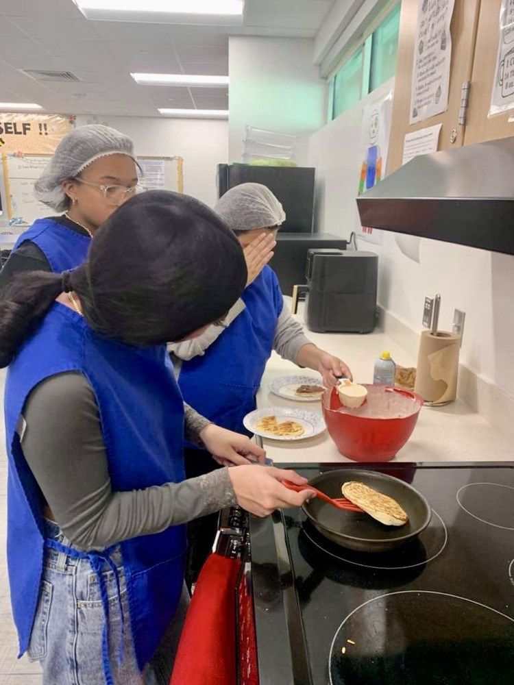 Alt text: Culinary students wearing aprons and hairnets proudly holding plates of freshly made pancakes and soft pretzels in a school kitchen.