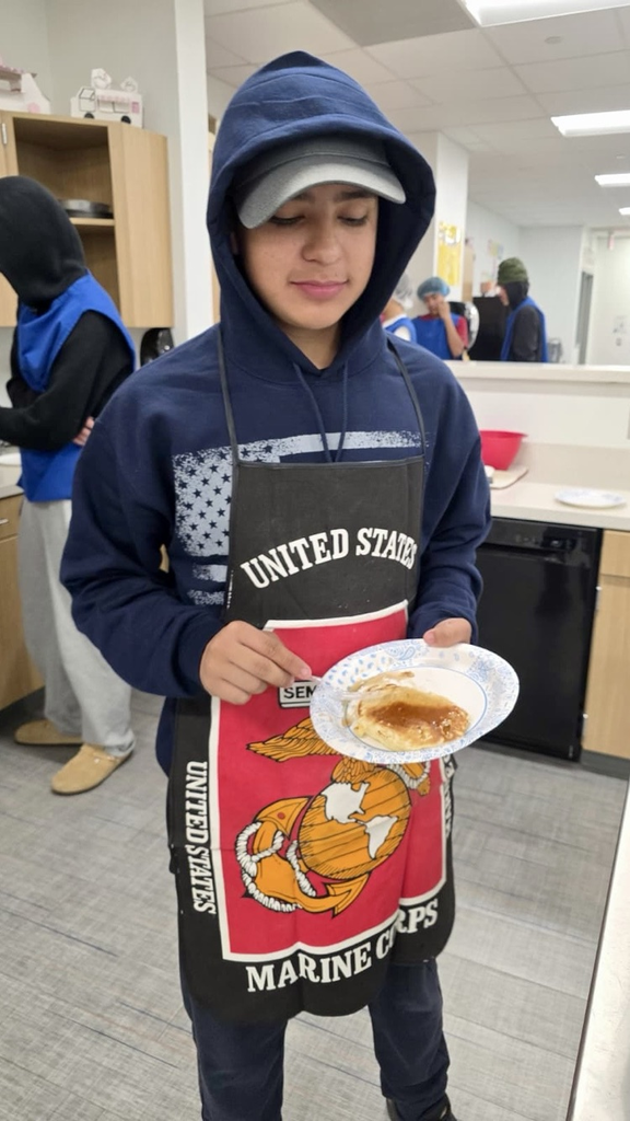 Alt text: Culinary students wearing aprons and hairnets proudly holding plates of freshly made pancakes and soft pretzels in a school kitchen.