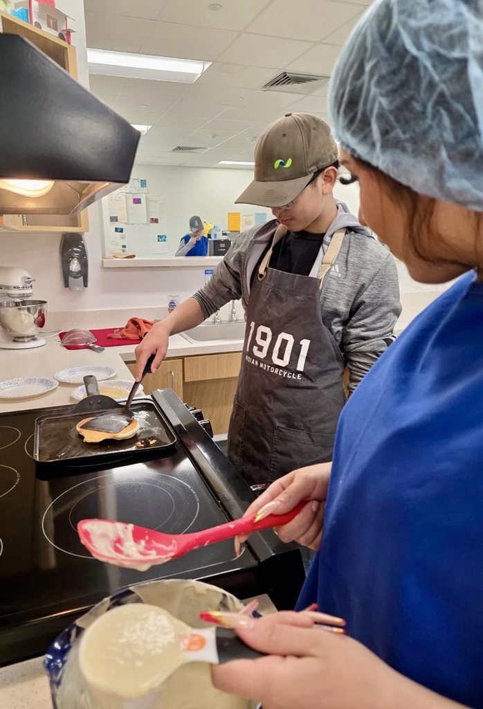 Alt text: Culinary students wearing aprons and hairnets proudly holding plates of freshly made pancakes and soft pretzels in a school kitchen.