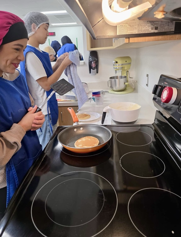 Alt text: Culinary students wearing aprons and hairnets proudly holding plates of freshly made pancakes and soft pretzels in a school kitchen.