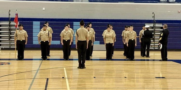 Alt text: NJROTC cadets in formal uniforms standing in formation inside a school gym during the Area Managers Inspection, including close-up and wide shots showing cadets at attention and leadership addressing the group.