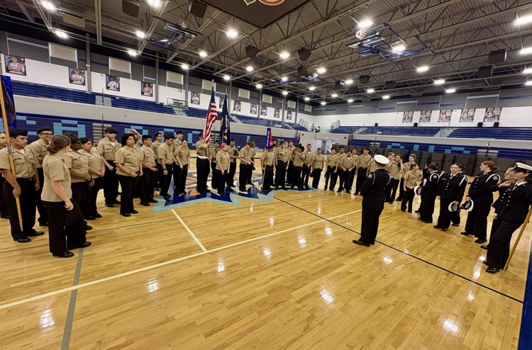 Alt text: NJROTC cadets in formal uniforms standing in formation inside a school gym during the Area Managers Inspection, including close-up and wide shots showing cadets at attention and leadership addressing the group.