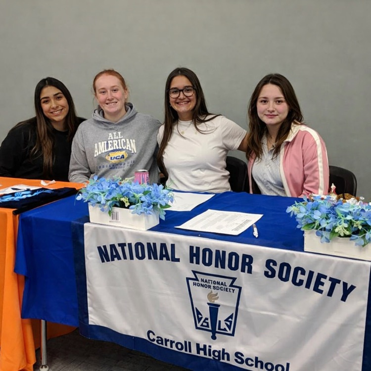  Four National Honor Society students sit behind a recruitment table during Tiger Time, smiling and posing in front of an NHS banner at Carroll High School with sign-up materials displayed on the table.