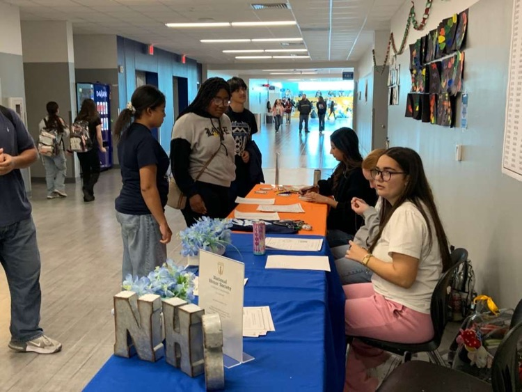  Four National Honor Society students sit behind a recruitment table during Tiger Time, smiling and posing in front of an NHS banner at Carroll High School with sign-up materials displayed on the table.