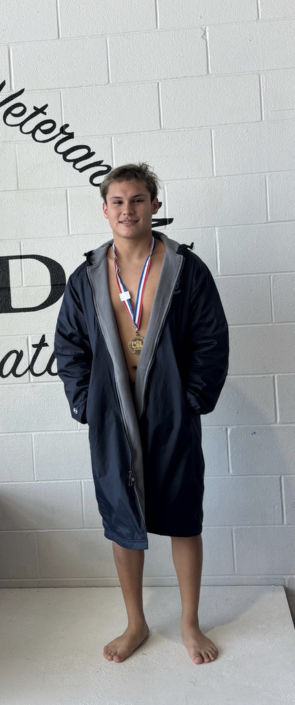 Alt Text: Student swimmer wearing a medal stands on the third-place podium at a swim meet, smiling, with a wall behind him displaying a UIL logo and aquatic center signage.