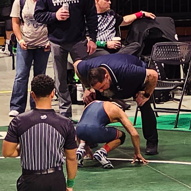 Carroll wrestlers kneeling together in a focused pre-match huddle on the blue mat at the state tournament.