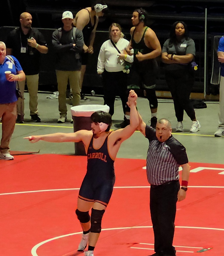 Carroll wrestlers kneeling together in a focused pre-match huddle on the blue mat at the state tournament.