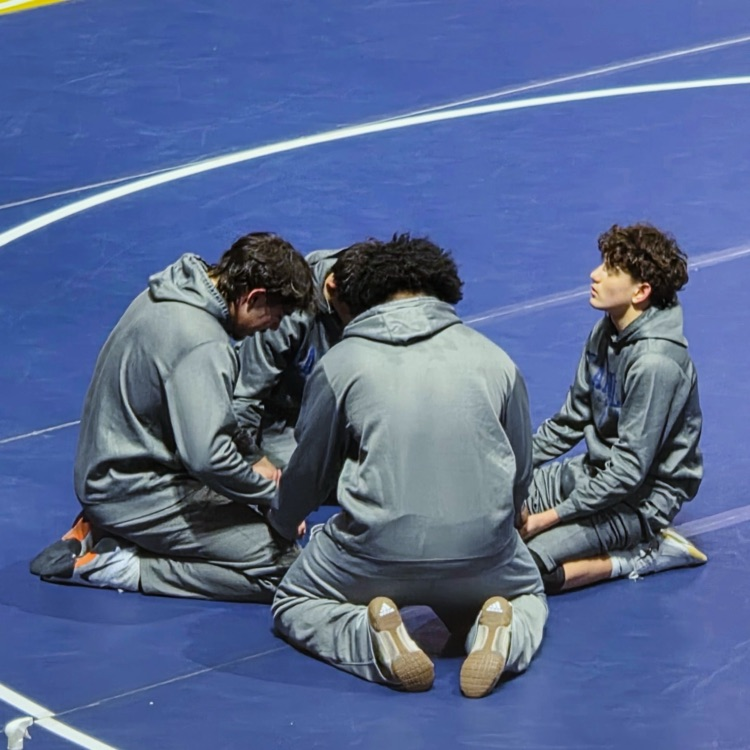 Carroll wrestlers kneeling together in a focused pre-match huddle on the blue mat at the state tournament.