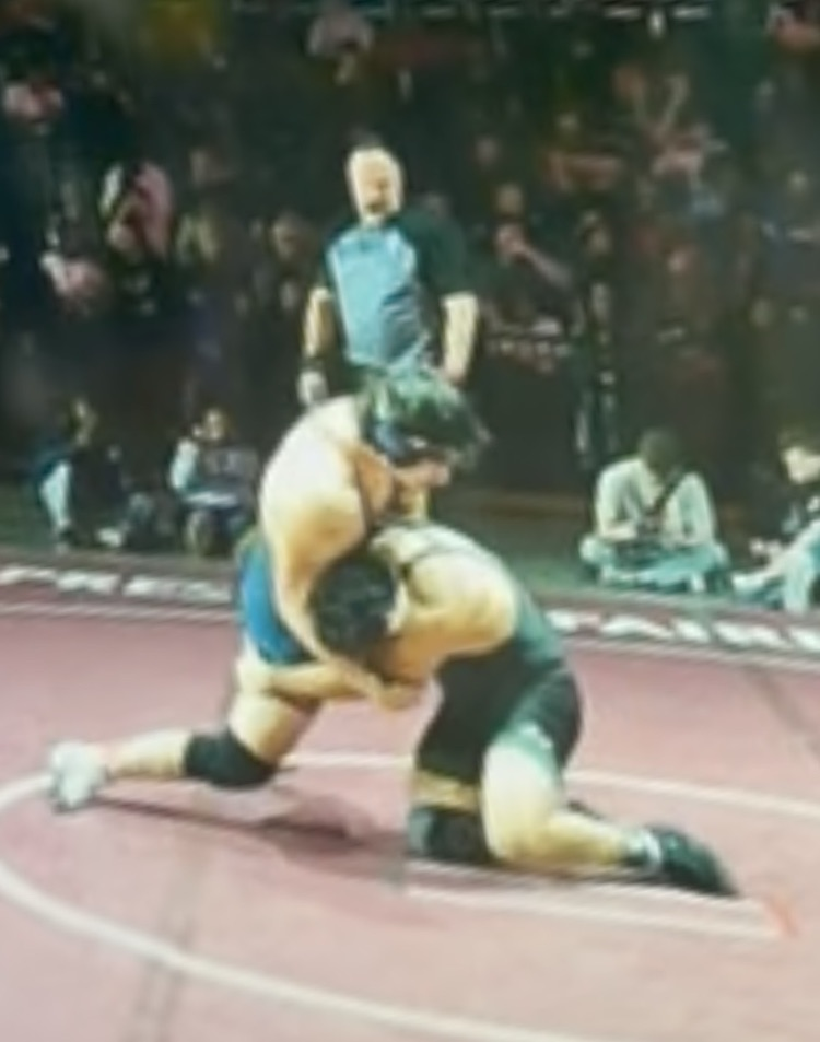 Carroll wrestler executes a takedown during a state championship match as the crowd watches in the background.