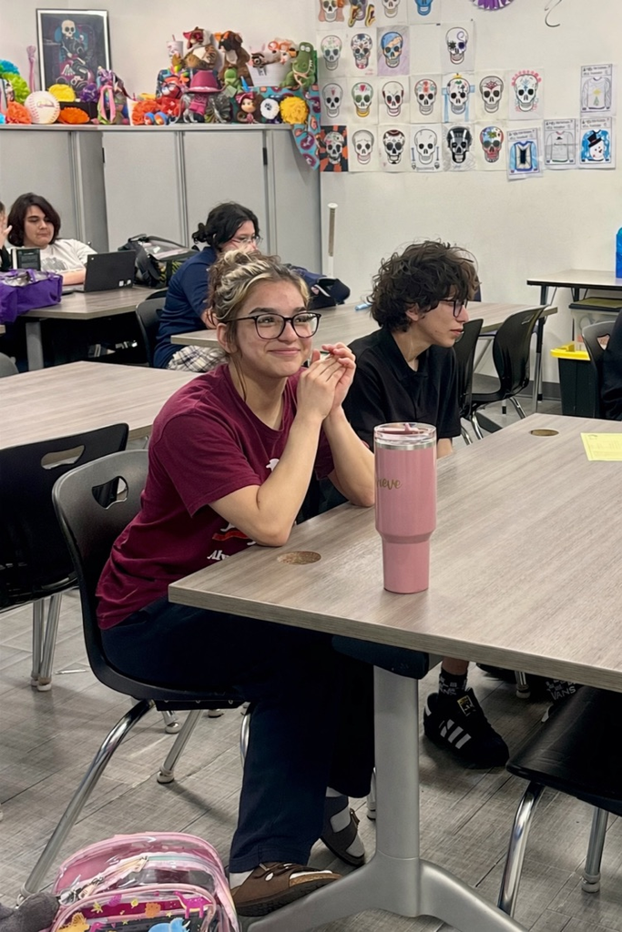 Student smiling at a classroom table with a pink tumbler, classmates working in the background.