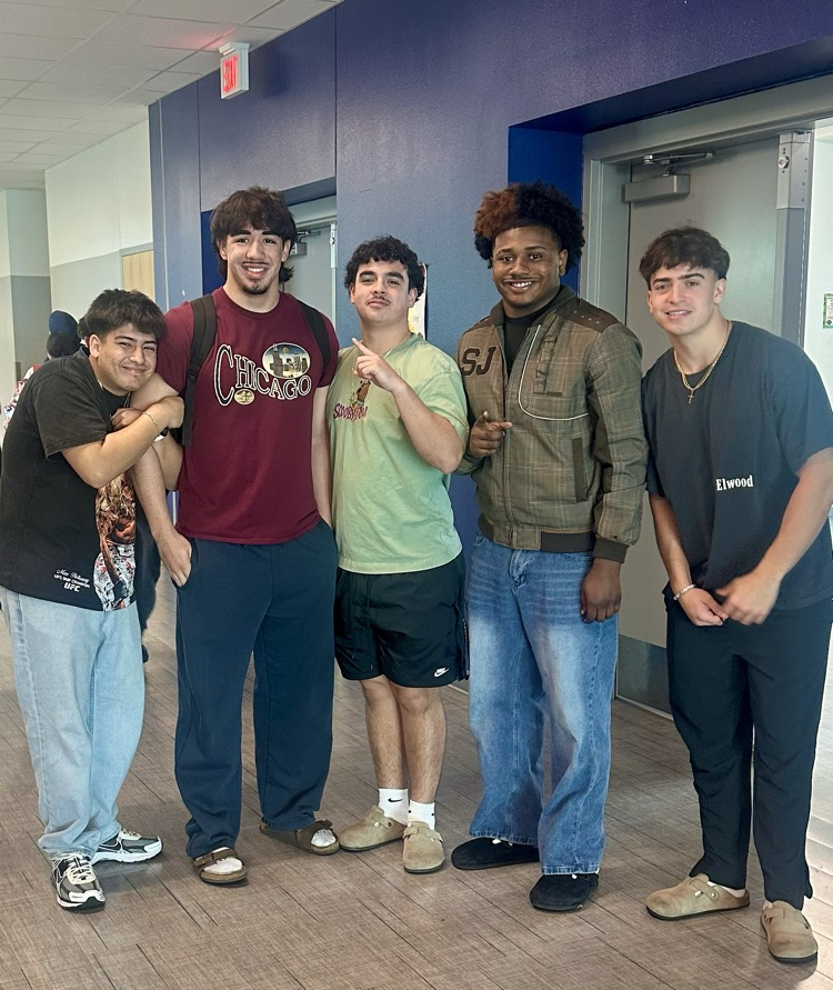 Student smiling at a classroom table with a pink tumbler, classmates working in the background.