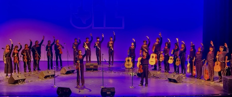 Mariachi students in black uniforms with red bow ties pose on a staircase holding a championship plaque, standing beside their director.